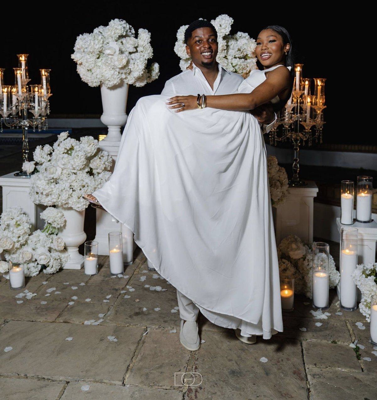 Denzil carrying Noelyne among candles and white flowers